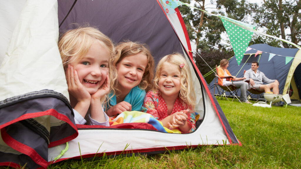 three girls in tent with parents on camping chairs in background
