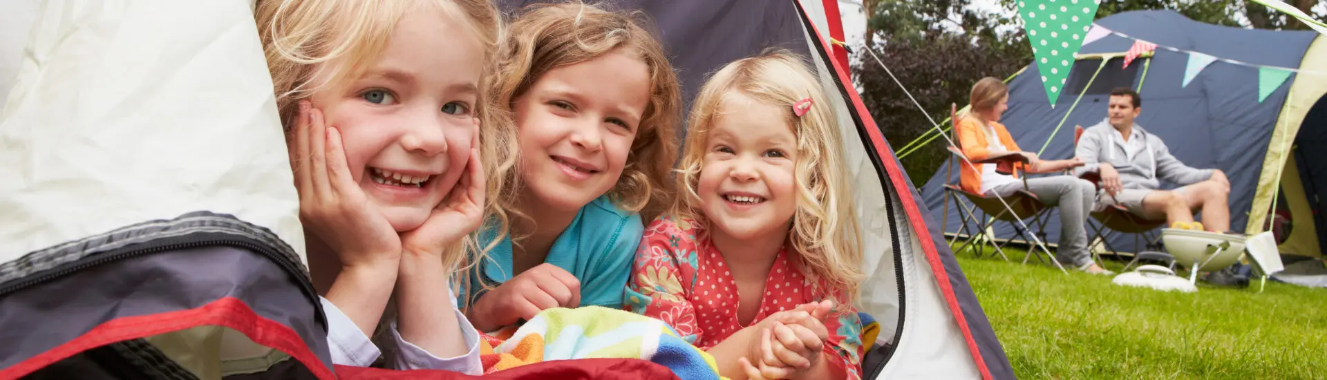three girls in tent with parents on camping chairs in background